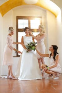 Eine Braut in einem weißen Kleid steht mit einem Blumenstrauß da, umgeben von drei Frauen, die ihr beim Frisieren und Anziehen helfen. Sie befinden sich in einem hellen Raum mit gefliestem Boden und einer gewölbten gelben Decke, und die Luft ist erfüllt von der Vorfreude auf die Hochzeit.