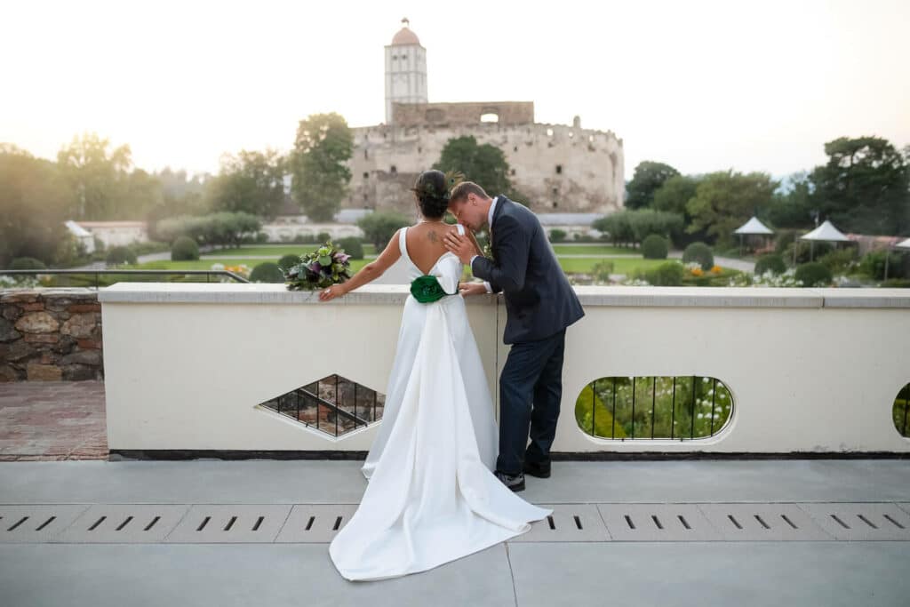 Eine Braut in einem weißen Kleid mit einem Blumenstrauß und ein Bräutigam im Anzug verbringen einen Moment ihrer Hochzeit auf einer Terrasse mit Blick auf ein historisches Steingebäude und einen Garten. Das Rückentattoo der Braut ist sichtbar, während der Bräutigam sie sanft auf die Schulter küsst und so ihre intime Hochzeitsfeier einfängt.