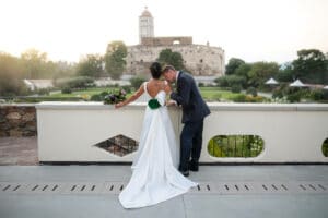 Eine Braut in einem weißen Kleid mit einem Blumenstrauß und ein Bräutigam im Anzug verbringen einen Moment ihrer Hochzeit auf einer Terrasse mit Blick auf ein historisches Steingebäude und einen Garten. Das Rückentattoo der Braut ist sichtbar, während der Bräutigam sie sanft auf die Schulter küsst und so ihre intime Hochzeitsfeier einfängt.