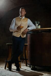 Ein Mann in goldener Weste und weißem Hemd lehnt an einem runden Stehtisch und hält ein Glas in der Hand. Die Kulisse scheint ein schwach beleuchteter, rustikaler Raum zu sein – perfekt für eine intime Hochzeitsplanung. Zwei Flaschen stehen auf dem Tisch neben ihm und tragen zur festlichen Atmosphäre bei.