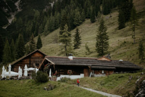 Eine rustikale Holzhütte, umgeben von einem bewaldeten Hügel, bietet die perfekte Kulisse für eine unvergessliche Hochzeit. Eine Reihe geschlossener weißer Sonnenschirme steht neben dem Gebäude, während eine Person in einer roten Jacke den schmalen Pfad durch die üppig grüne Landschaft entlangläuft und Ihrer Hochzeitskulisse Charme verleiht.