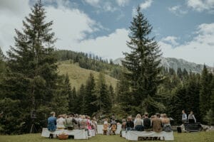Eine malerische Hochzeitszeremonie im Freien vor der Kulisse üppiger grüner Berge und hoher Kiefern. Die Gäste sitzen auf weißen Bänken und der Himmel ist teilweise bewölkt, was die perfekte Atmosphäre zum Heiraten schafft.