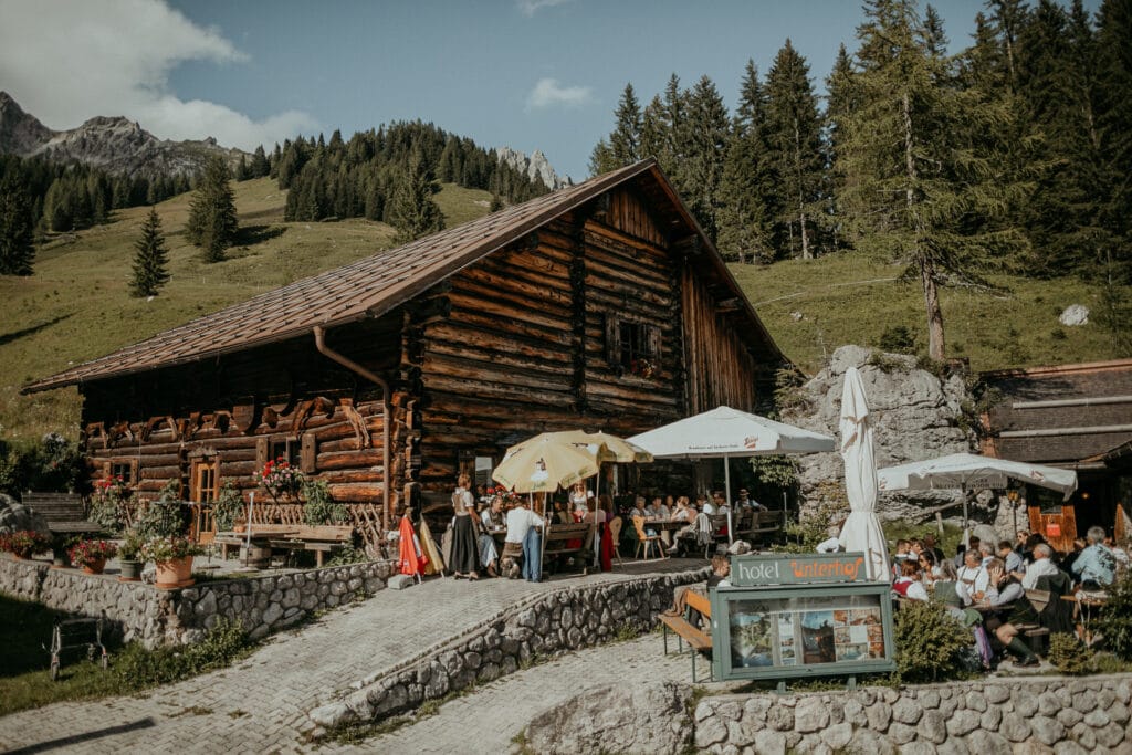 Eine rustikale Holzhütte in einer Bergregion bietet mit üppigen Bäumen und einer grasbewachsenen Landschaft die perfekte Kulisse für Heiraten. Die Gäste sitzen unter Sonnenschirmen an Tischen im Freien und genießen die malerische Umgebung. Ein charmanter Weg führt zum Eingang der Hütte.