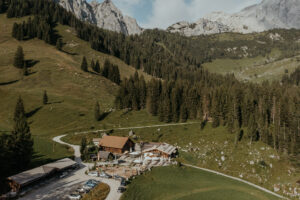 Eine Luftaufnahme einer rustikalen Lodge, eingebettet in ein üppiges grünes Tal, umgeben von dichten Kiefernwäldern und felsigen Bergen, perfekt für eine Hochzeit. Mehrere Autos sind in der Nähe der Lodge geparkt und deuten auf laufende Hochzeitsplanungen hin, während eine kurvenreiche Straße elegant durch die bezaubernde Landschaft führt.