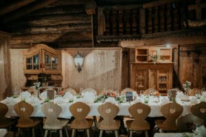 Ein rustikales Esszimmer, perfekt für eine Hochzeitsplanung, mit einem langen Holztisch, der für ein Essen gedeckt ist. Weiße Tischdecken, Servietten, Glaswaren und Blumengestecke schmücken das Ambiente. Holzstühle mit herzförmigen Ausschnitten umgeben den Tisch und ergänzen die Holzwände und -balken wunderschön.