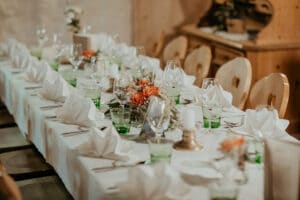 Ein langer Esstisch ist elegant für eine Hochzeit gedeckt, mit weißen Tischdecken, ordentlich gefalteten Servietten und grünen Gläsern. Blumengestecke mit orangefarbenen und weißen Blumen schmücken den Tisch, umgeben von polierten Holzstühlen in einer gemütlichen Umgebung, die perfekt für Hochzeiten ist.