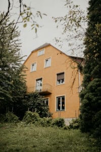 Inmitten üppigen Grüns und hoher Bäume bietet dieses charmante dreistöckige, orangefarbene Haus mit kleinem Balkon eine malerische Kulisse für alle, die von einer Hochzeitsfeier unter bewölktem Himmel träumen. Mit mehreren Fenstern, die für natürliches Licht sorgen, ist es der perfekte Ort für Ihren romantischen Hochzeitstag.