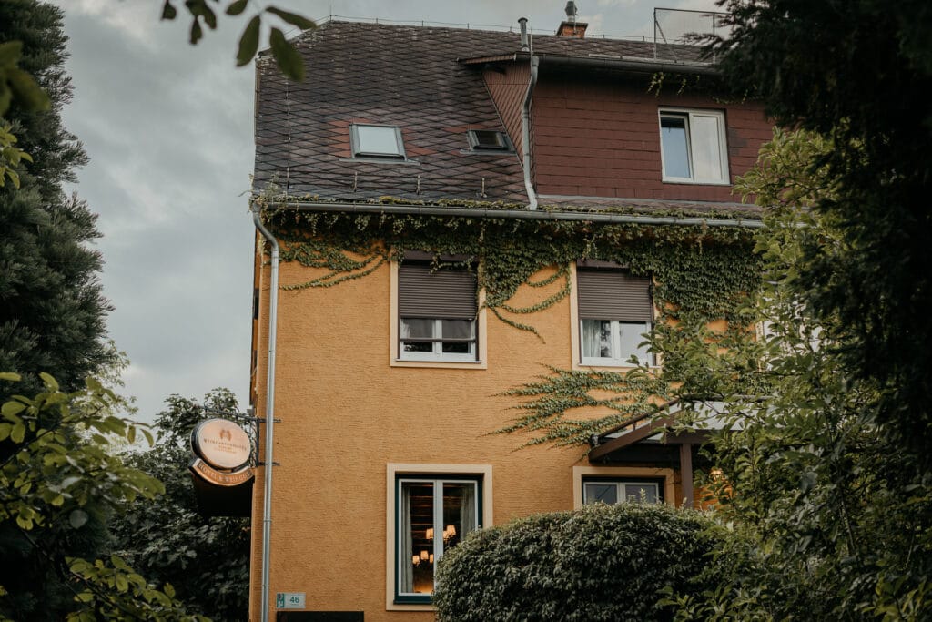 Ein charmantes zweistöckiges Haus mit einem schrägen, mit Efeu bewachsenen Dach, gelben Wänden und braunen Akzenten. Ein rundes Schild hängt in der Nähe des Eingangs, umgeben von üppigem Grün und Büschen, und bietet eine malerische Kulisse für alle, die von einer romantischen Hochzeit unter bewölktem Himmel träumen.