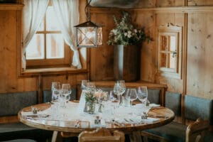 Ein gemütliches, holzgetäfeltes Esszimmer mit einem runden Tisch, der wunderschön für ein Essen gedeckt ist, perfekt für eine intime Hochzeitsfeier. Der Tisch ist mit elegantem Glasgeschirr, Blumenarrangements und ordentlich angeordnetem Besteck geschmückt. Darüber hängt eine warme Hängelampe, die ein sanftes Licht verbreitet.