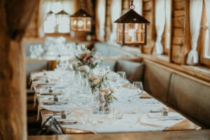 Ein gemütliches Restaurant mit einem langen Holztisch, der perfekt für eine intime Hochzeit gedeckt ist. Elegantes Glasgeschirr, gefaltete Servietten und Blumengestecke sind sorgfältig arrangiert. Warme Hängelampen und sanftes Tageslicht schaffen eine einladende Atmosphäre, die ideal für Ihre Hochzeitsplanung ist.