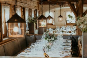 Ein rustikales Restaurant mit einem langen, elegant gedeckten Tisch, perfekt für Hochzeitsfeiern. Geschmückt mit Blumen, Gläsern und Besteck sorgt das warme Licht der hängenden Laternen für eine gemütliche Atmosphäre. Holzbalken und große Fenster verstärken den Charme.