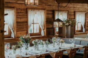 Rustikaler Speisesaal mit Holzinterieur, perfekt für eine Hochzeit, mit einem langen Tisch, geschmückt mit weißen Tischdecken, Blumengestecken und Glaswaren. Sanftes Tageslicht strömt durch die Fenster mit Vorhängen und schafft eine warme und einladende Atmosphäre, ideal für die Hochzeitsplanung.