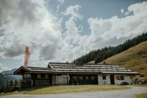 Eine rustikale Holzhütte mit schrägem Dach steht auf einem grasbewachsenen Hügel unter einem teilweise bewölkten Himmel, perfekt für eine intime Hochzeitsatmosphäre. Ein hoher Fahnenmast mit einer rot-weißen Flagge steht in der Nähe, während Kiefern eine natürliche Kulisse für unvergessliche Hochzeitsplanungsmomente schaffen.