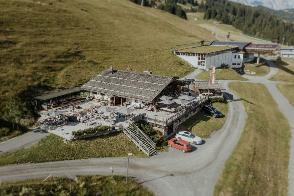 Luftaufnahme eines rustikalen Bergrestaurants mit Holzdach, perfekt für alle, die eine Hochzeitsfeier planen. Umgeben von grasbewachsenen Hügeln sind mehrere Autos in der Nähe geparkt und Gäste sitzen an Tischen im Freien. Im Hintergrund sind Gebäude zu sehen und gewundene Straßen bieten idyllische Ausblicke.