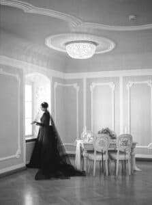 Eine Frau in einem eleganten schwarzen Kleid und Schleier steht an einem Fenster in einem wunderschön dekorierten Raum, der an eine Szene aus einer sorgfältigen Hochzeitsplanung erinnert. Der Raum verfügt über aufwendige Wandleisten, einen Kronleuchter und einen gedeckten Esstisch mit Stühlen, Kerzen und Blumenarrangements. Die Stimmung ist heiter und kultiviert.