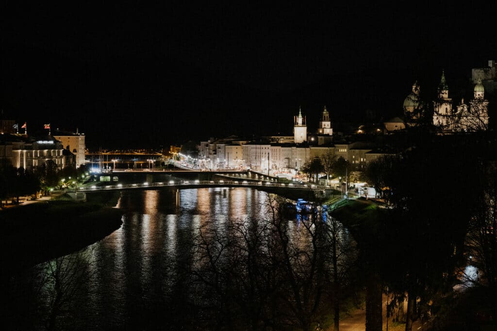 Eine nächtliche Ansicht einer Stadt am Fluss, perfekt für eine Hochzeit, beleuchtet durch Lichter, die sich im Wasser spiegeln. Eine Brücke verbindet die beiden Flussufer, und Gebäude mit beleuchteten Fenstern säumen die Uferpromenade. Eine schwache Silhouette eines Hügels schafft eine romantische Kulisse, ideal für die Hochzeitsplanung.