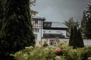 Ein charmantes Gebäude mit der Aufschrift „Parkhotel“, umgeben von üppigem Grün und hohen Bäumen, bietet eine idyllische Kulisse für Ihre Hochzeit. Die Szene spielt unter einem bewölkten Himmel und schafft eine ruhige Atmosphäre mit blühenden Büschen im Vordergrund – perfekt für die Planung Ihrer Traumhochzeit.