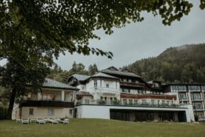Ein malerischer Blick auf ein großes, elegantes Gebäude, eingebettet in eine üppige, grüne Landschaft, ist die perfekte Kulisse für eine Hochzeit. Das Gebäude verfügt über mehrere Ebenen, Balkone und elegante Holzelemente. Im Vordergrund befindet sich eine Rasenfläche mit Liegestühlen, eingerahmt von Ästen – ideal für die Hochzeitsplanung.
