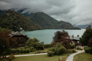 Eine ruhige Landschaft, perfekt für eine Hochzeit, mit einem klaren See, umgeben von üppigen, bewaldeten Bergen unter einem bewölkten Himmel. Im Vordergrund schaffen rustikale Holzhäuser und eine Rasenfläche entlang des gewundenen Pfads, der von Sträuchern und Bäumen gesäumt ist, eine idyllische Kulisse für die Hochzeitsplanung.