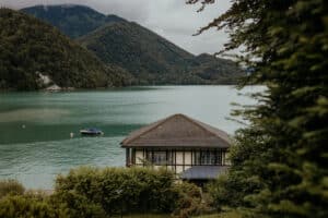 Malerische Aussicht auf einen ruhigen See, umgeben von üppigen grünen Hügeln, perfekt für eine traumhafte Hochzeit. Im Vordergrund steht ein kleines, rustikales Haus nahe dem Wasserrand und ein Boot schwimmt in der Nähe. Der bewölkte Himmel schafft eine ruhige und stimmungsvolle Atmosphäre, ideal für intime Hochzeitsmomente.