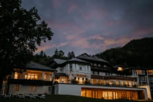 Ein großes, beleuchtetes Gebäude steht in der Abenddämmerung inmitten einer Berglandschaft, perfekt für eine Traumhochzeit. Das Gebäude verfügt über mehrere Fenster und Balkone, ist von Bäumen und grünen Hügeln umgeben und schafft eine zauberhafte Kulisse, um die Hochzeit unter dem dunkler werdenden Himmel zu beginnen.