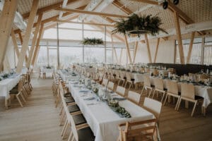 Ein geräumiger, hölzerner Bankettsaal ist elegant für eine Hochzeit eingerichtet, mit langen Tischen, die mit weißen Tischdecken bedeckt sind. Jeder Tisch ist mit Grünpflanzen und Glaswaren geschmückt. Große Fenster lassen natürliches Licht herein und bieten Ausblicke ins Freie – perfekt für Ihre Hochzeitsplanungsträume.
