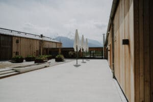 Ein modernes Gebäude mit Holzwänden und mehreren geschlossenen weißen Sonnenschirmen schmückt einen geräumigen Innenhof, der sich perfekt für Hochzeitsveranstaltungen eignet. Blumenbeete säumen den Weg, während in der dunstigen Ferne Berge unter einem wolkigen Himmel eine romantische Kulisse bilden.