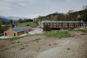 Eine malerische ländliche Landschaft mit modernen Holzgebäuden, ideal für eine Hochzeit, umgeben von grünen Hügeln und fernen Bergen unter einem bewölkten Himmel. Das zeitgenössische Design mit großen Fenstern harmoniert perfekt mit der ruhigen Umgebung und bietet eine malerische Szene für diejenigen, die ihren besonderen Tag planen.