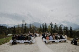 Eine Hochzeitszeremonie findet im Freien statt, wobei ein großes Publikum vor einem Blumenbogen sitzt. Mit der Magie der Hochzeitsplanung werden üppige Bäume und majestätische, schneebedeckte Berge unter einem bewölkten Himmel als Kulisse geschaffen.