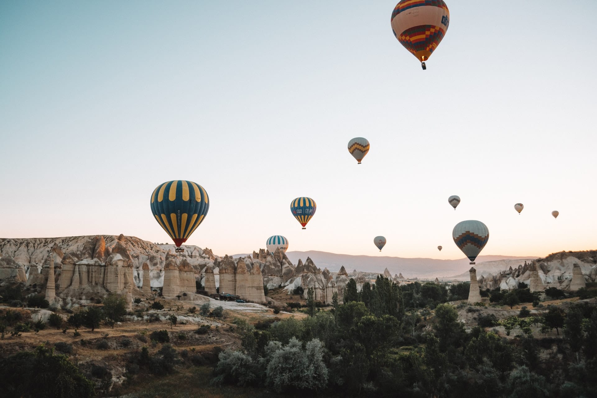 Heißluftballons schweben bei Sonnenaufgang über einer felsigen Landschaft und schaffen eine traumhafte Kulisse, die perfekt für eine märchenhafte Hochzeit ist. Einzigartige Felsformationen und viel Grün bereichern die ruhige, malerische Szene, ideal für eine unvergessliche Hochzeitsplanung unter einem klaren, hellen Himmel.