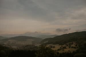 Eine ruhige Hügellandschaft mit üppigem Grün unter einem wolkigen Himmel bildet die perfekte Kulisse für eine romantische Hochzeit. Die Berge in der Ferne und die verstreuten Bäume schaffen eine friedliche und natürliche Szene, ideal für unvergessliche Hochzeitsplanungsmomente.