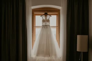 Ein weißes Brautkleid mit Spitzendetails am Mieder hängt vor einem Fenster mit Holzrahmen und spiegelt die Essenz von Hochzeit wider. Der Raum ist sanft beleuchtet, dunkle Vorhänge sind teilweise zugezogen und rechts steht eine Stehlampe.