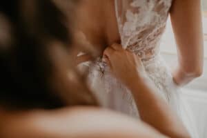 Nahaufnahme einer Person, die einer anderen Person hilft, ein Spitzenhochzeitskleid zuzuknöpfen, wobei die aufwendigen Details hervorgehoben werden. Dieser zarte und intime Moment fängt die Essenz des Heiratens ein, bei dem jeder sorgfältig geschlossene Knopf Teil wertvoller Erinnerungen wird.