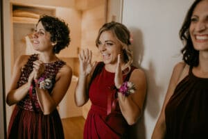 Drei Frauen in burgunderfarbenen Kleidern reagieren freudig in einem warm beleuchteten Raum, möglicherweise während eines besonderen Anlasses wie einer Hochzeit. Mit ihren Ansteckblumen wirken sie aufgeregt und glücklich, vielleicht gefangen im glückseligen Wirbelwind der Hochzeitsplanung.