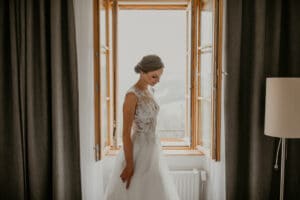 Eine Braut in einem eleganten weißen Brautkleid steht an einem offenen Fenster, das sanft von natürlichem Licht erhellt wird. Während sie in Gedanken an ihre bevorstehende Hochzeit versunken ist, bilden die dunklen Vorhänge einen Kontrast zur hellen Szene. Sie blickt mit einem sanften Lächeln nach unten, während neben ihr eine Stehlampe zu sehen ist.