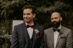 Zwei Männer in Anzügen stehen draußen, einer mit roter Fliege und Boutonniere lächelnd, der andere mit Bart und dunkler Fliege sieht zufrieden aus. Sie stehen vor üppigem Grün und schwelgen vielleicht in der Freude ihrer Hochzeit.
