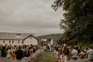 Eine Hochzeit findet im Freien statt, während die Gäste auf beiden Seiten eines blumengesäumten Ganges sitzen. Das Paar steht am Altar, eingerahmt von einem rustikalen Gebäude und üppigen grünen Hügeln unter einem wolkigen Himmel, alles perfekt zusammengefügt durch sorgfältige Hochzeitsplanung.