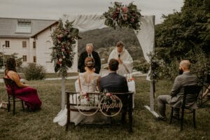 Ein Paar sitzt unter einem mit Blumen geschmückten Hochzeitsbogen im Freien und genießt die Schönheit seiner Hochzeit. Der in Weiß gekleidete Geistliche steht vor ihnen. Zwei Gäste in formeller Kleidung sitzen auf beiden Seiten, während ein Gebäude und üppige grüne Hügel eine atemberaubende Kulisse für diese sorgfältig orchestrierte Hochzeitsplanung bieten.