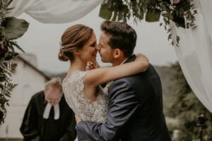 In freudiger Umarmung unter dem elegant dekorierten Bogen feiern Braut und Bräutigam ihre Hochzeit. In einem dunklen Anzug und einem Spitzenkleid stehen sie vereint da, während im Hintergrund jemand aus einem Buch vorliest, was ihre Hochzeitszeremonie im Freien wirklich unvergesslich macht.