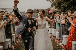 Ein fröhliches Paar schreitet durch einen mit Rosenblättern geschmückten Gang im Freien. Der Bräutigam reckt jubelnd die Faust in die Höhe, während die Braut einen Blumenstrauß hält. Die bunt gekleideten Gäste jubeln und pusten Seifenblasen um sie herum – ein perfekter Moment, eingefangen in ihrer gut durchdachten Hochzeitsplanung.
