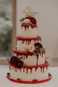 Eine vierstöckige weiße Torte mit rotem Zuckerguss und Blumendekorationen, darunter rosa und dunkelrote Blumen, steht elegant auf einem roten Tablett. Perfekt für eine Hochzeit, auf der Torte steht „Lasst das Abenteuer beginnen“ und gibt damit den Ton für Ihre Hochzeitsreise an.