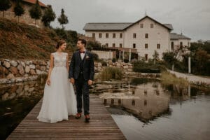 Braut und Bräutigam halten sich auf einem Holzsteg neben einem ruhigen Teich an den Händen, ihr Spiegelbild spiegelt sich im Wasser. Die Planung dieser Hochzeit war perfekt; sie trägt ein weißes Kleid, während er einen dunklen Anzug trägt. Unter einem bewölkten Himmel vervollständigen ein großes Haus und üppiges Grün die malerische Kulisse für ihre Hochzeit.