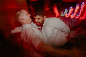 Zwei Männer in weißen Hemden tanzen fröhlich zusammen in einem schwach beleuchteten Raum mit verschwommenen Neonlichtern im Hintergrund und schaffen so eine lebhafte und festliche Hochzeitsatmosphäre.