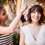 Eine Frau in einem weißen Spitzenkleid lächelt, während eine andere Frau, vielleicht ein Mitglied ihres Hochzeitsplanungsteams, ihren Blumenkopfschmuck zurechtrückt. Sie stehen draußen, umgeben von üppigem Grün und vor einer bezaubernden gelben Wand, die die Freude des Heiratens einfängt.