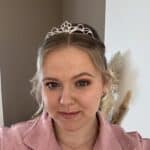 Eine Frau mit blondem Haar, die ein zierliches Diadem und eine rosafarbene strukturierte Bluse trägt, lächelt sanft in die Kamera. Im Hintergrund sind eine zweifarbige Wand und einige dekorative getrocknete Gräser zu sehen, die die Kunstfertigkeit der Maskenbildnerin Katharina Rosner unterstreichen.