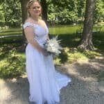 Eine Braut in einem weißen Spitzenhochzeitskleid hält einen Strauß weißer Blumen und Grünzeug in der Hand, während sie auf einem Kiesweg in einem sonnenbeschienenen Park steht. Ihr strahlendes Aussehen wurde von der Visagistin Katharina Rosner wunderschön in Szene gesetzt.