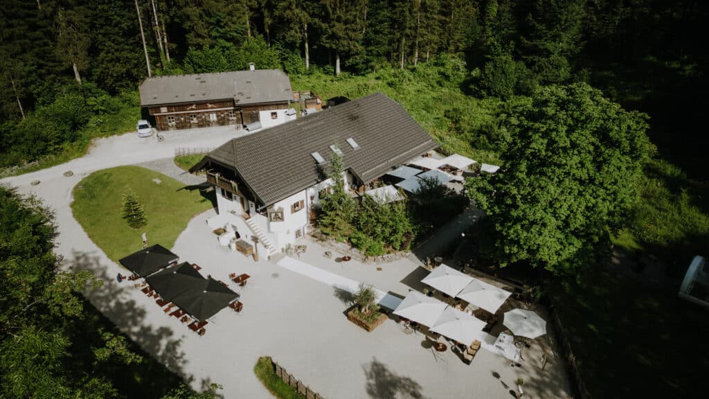 Luftaufnahme eines rustikalen Hauses, das sich ideal für Hochzeitsfeiern eignet und von üppigem Grün und Wald umgeben ist. Das Anwesen verfügt über Sitzgelegenheiten im Freien mit Sonnenschirmen und einen Kiesweg, der zum Eingang führt. In der Nähe sind ein Nebengebäude und ein geparktes Fahrzeug zu sehen. Perfekt für diejenigen, die inmitten der Natur heiraten möchten.