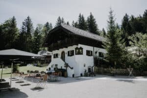 Ein malerisches europäisches Cottage mit Holzdach und weißen Wänden liegt eingebettet in einem ruhigen Wald und ist perfekt für eine intime Hochzeit. Auf der Kiesterrasse, umgeben von hohen Kiefern unter einem klaren blauen Himmel, erwarten Sie hölzerne Tische und Stühle für den Außenbereich.