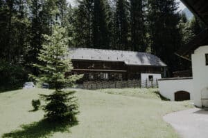 Ein rustikales Holzhaus mit grauem Dach liegt eingebettet zwischen hohen immergrünen Bäumen, perfekt für alle, die von einer romantischen Hochzeit träumen. Ein kleines weißes Auto ist in der Nähe geparkt. Ein abschüssiger Rasen mit einer einzelnen Kiefer führt zum Haus, während ein Holzzaun um das Grundstück verläuft.