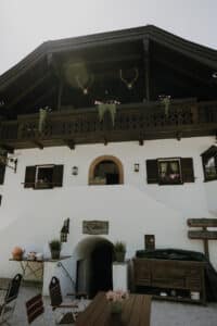 Ein rustikales Alpenhaus, perfekt für eine Hochzeit, verfügt über einen dunklen Holzbalkon, der mit Geweihen und Topfpflanzen geschmückt ist. Darunter ergänzt eine weiße Fassade mit braunen Fensterläden die Gartenmöbel und Schilder. Ein paar Dekorationen zieren den Tisch und schaffen eine malerische und charmante Umgebung, die ideal zum Heiraten ist.